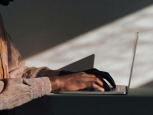 A close-up side view of a person's hands typing on a silver laptop on a desk, with dramatic shadows cast across the background.